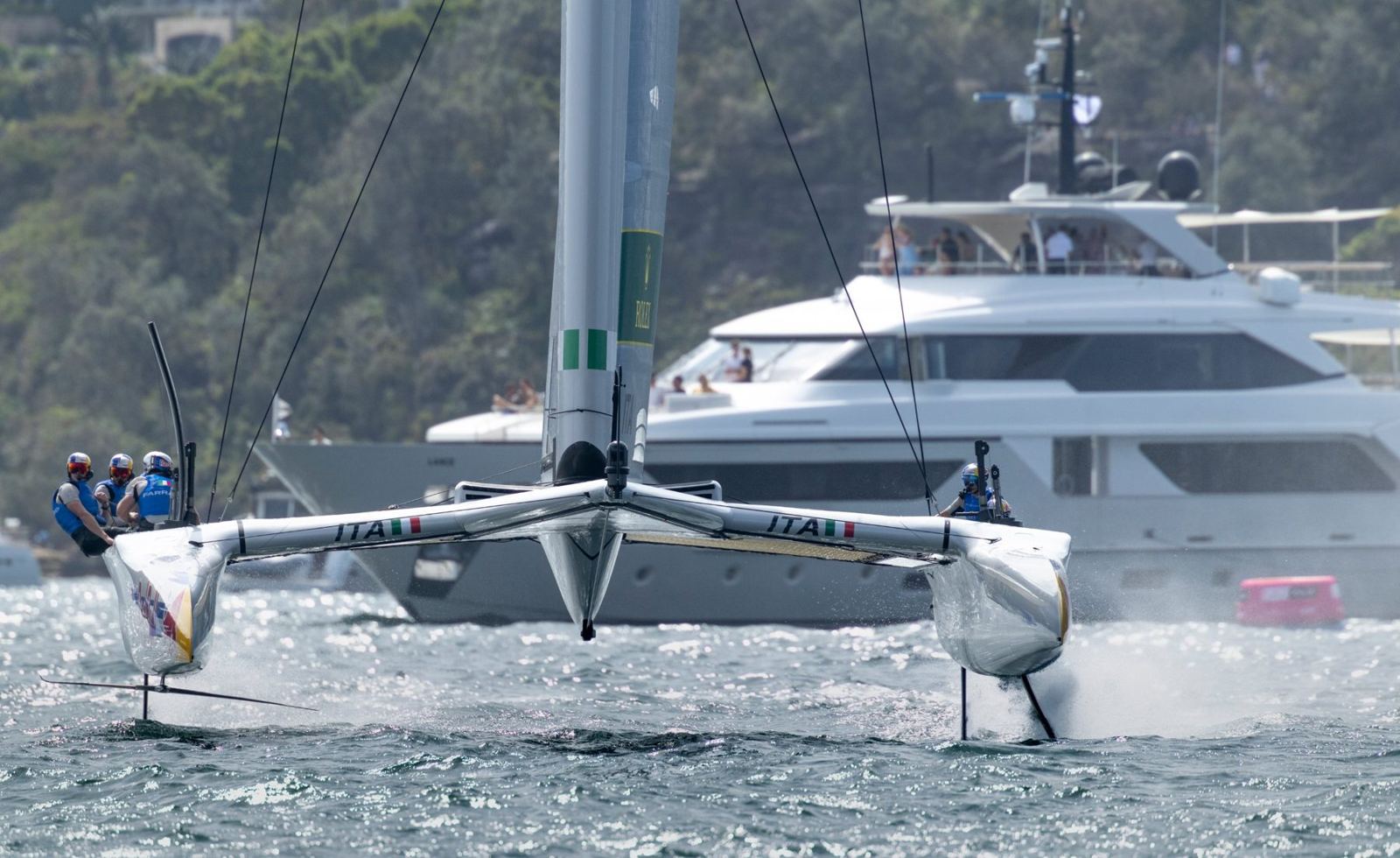 An F50 catamaran foiling past a superyacht with guests on deck