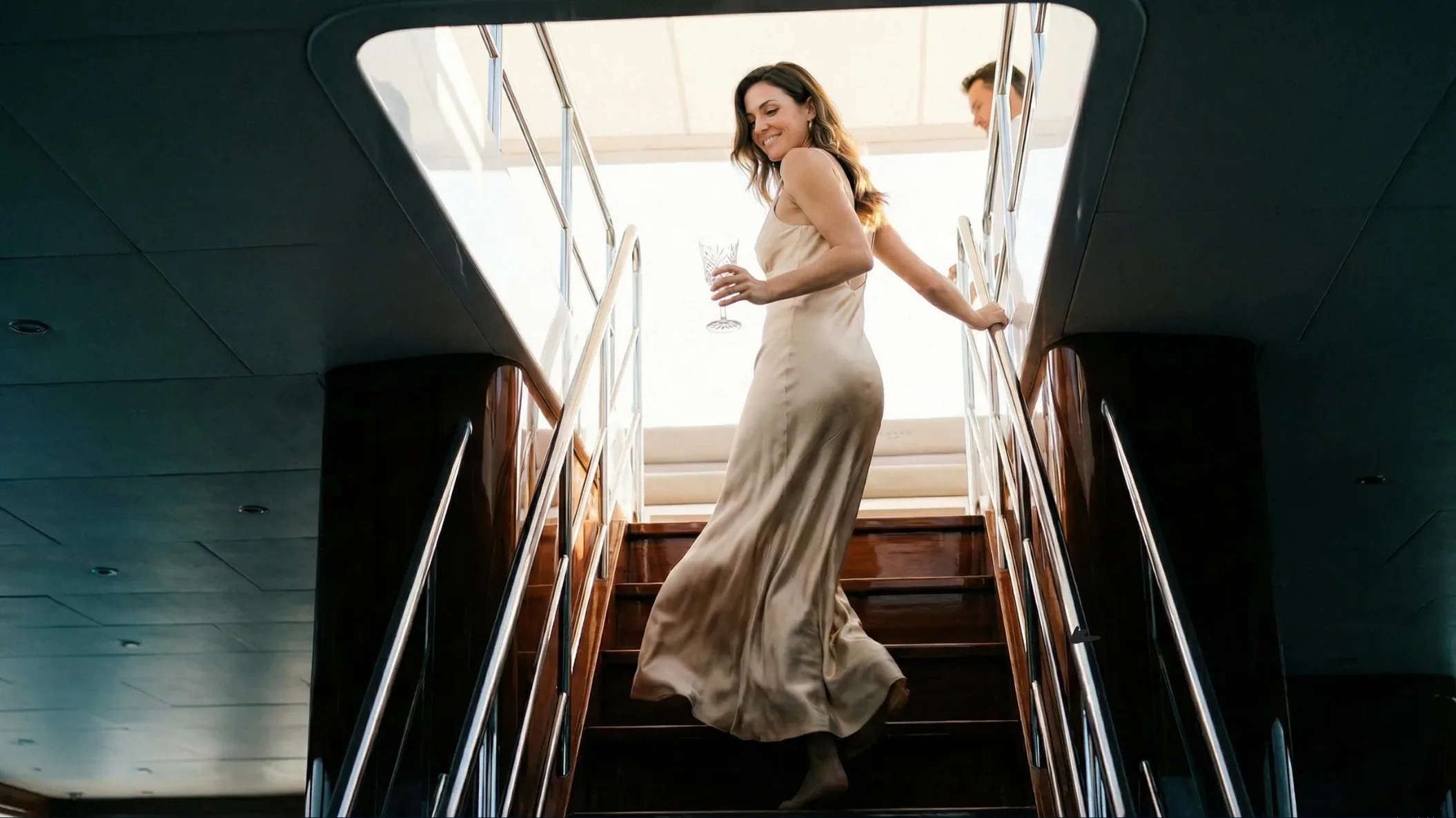 A woman in a satin dress walking down the stairs of a yacht with a glass of champagne